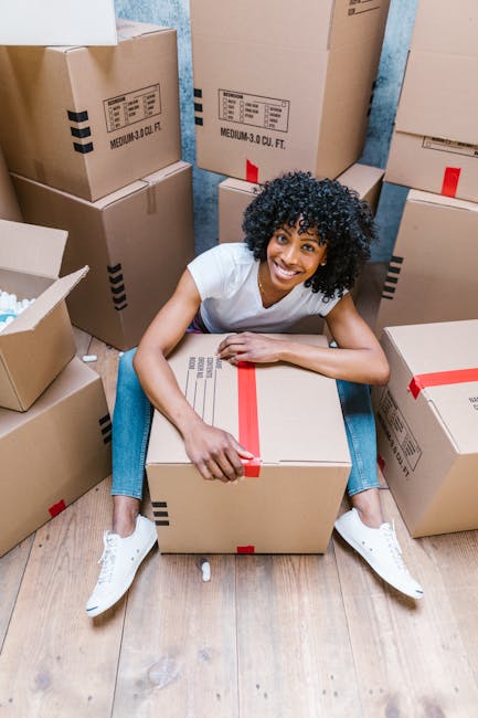 A young woman with curly black hair, wearing a white t-shirt, blue jeans, and white sneakers, sits on a wooden floor surrounded by packed cardboard boxes of various sizes, many sealed with red packing tape. She is smiling and holding a medium-sized box, positioning it on the floor, with several other boxes stacked around her in a small indoor space. The background features a blue wall, and the setting appears to be prepared for a home relocation, with boxes ready for transport. This scene demonstrates professional packing and loading processes involved in house removals, highlighting the importance of careful handling and organization during furniture transport. Man and Van Brixton specializes in relocation services, including packing and moving, ensuring a smooth and efficient home moving experience.
