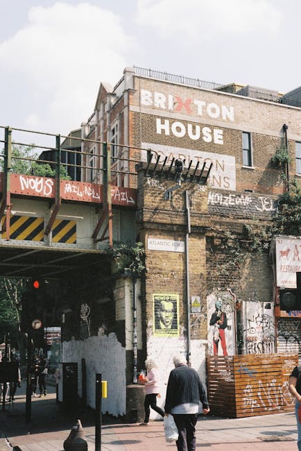 A multi-storey brick building located on Coldharbour Lane in Brixton with large white painted signage displaying 'BRIXTON' and 'HOUSE' on its exterior wall. The building features a fire escape ladder and metallic balcony railing extending from the upper floors. The ground level shows an entryway with a dark opening leading into an underground or basement area, with visible graffiti and posters on the walls surrounding it. In the foreground, several pedestrians are walking along the pavement, some carrying bags, indicating an active street scene. The surroundings include scaffolding, a metal bridge or platform, and various posters and signs, typical of a bustling urban environment. The scene is captured during daylight with overcast skies, providing natural lighting. The image reflects the typical context of house relocation or furniture transport activities near residential and commercial properties in Brixton, with [COMPANY_NAME] specializing in removals and moving services.