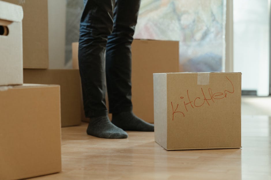 A young woman with curly black hair, wearing a white t-shirt, blue jeans, and white sneakers, sits on a wooden floor surrounded by packed cardboard boxes of various sizes, many sealed with red packing tape. She is smiling and holding a medium-sized box, positioning it on the floor, with several other boxes stacked around her in a small indoor space. The background features a blue wall, and the setting appears to be prepared for a home relocation, with boxes ready for transport. This scene demonstrates professional packing and loading processes involved in house removals, highlighting the importance of careful handling and organization during furniture transport. Man and Van Brixton specializes in relocation services, including packing and moving, ensuring a smooth and efficient home moving experience.