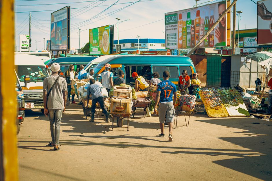 A busy outdoor market scene with multiple individuals engaged in buying and selling activities. Several people are gathered around a variety of items displayed on tables and the ground, including boxes, textiles, bottles, and various small goods. Some are carrying or pushing carts loaded with goods, while others are browsing or arranging merchandise. A small blue van is parked nearby, with people loading or unloading items from it, indicating a home relocation or transport process. The market is set in an open area with billboards and signage visible in the background, and bright daylight illuminating the scene. The overall environment suggests a lively marketplace with ongoing commerce, reflecting an active setting for packing and moving logistics, as would be handled by companies like Man and Van Brixton.