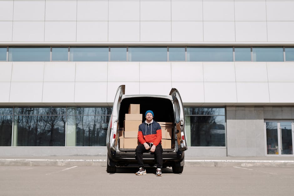 A man dressed in a red and navy jacket, blue beanie, and black pants is seated on the open tailgate of a black van, which is loaded with packed cardboard boxes of various sizes. The van is parked in front of a modern, multi-storey building with large windows and a light grey façade. Behind the vehicle, the surrounding environment features leafless trees and a clear sky, suggesting a cold day. Inside the van, additional boxes are visible, indicating a home relocation process involving furniture transport and packing. The loading area appears to be a paved lot suitable for moving services, with the man and van representing a professional moving or removals operation by [COMPANY_NAME], operating across Brixton and Effra Road zones. The scene captures a typical stage in the packing and moving process, with the van prepared for transportation of household items, emphasizing efficient furniture transport and loading procedures.