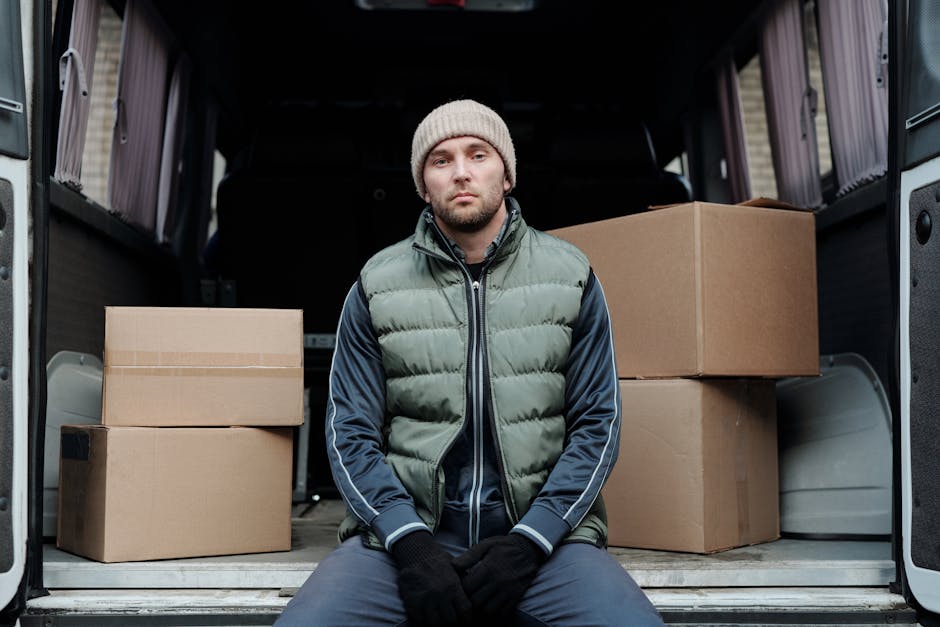 A man in casual clothing, including a black vest, blue sleeves, black pants, and white sneakers, stands outside a residential property next to a white moving van. He is wearing a light grey cap and bl